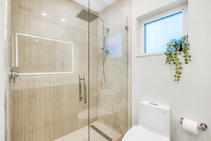 Bright modern bathroom with glass walk-in shower, wood-effect wall paneling and hexagon tile, rainfall showerhead, white toilet and small window with trailing plant