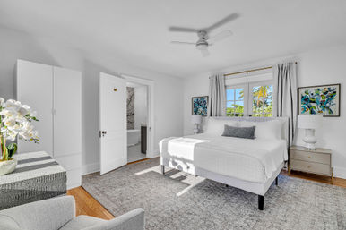 Bright sunlit coastal bedroom with king bed in white linens, gray rug, ceiling fan, orchids on a dresser, palm-tree view through the window and an ensuite bathroom.