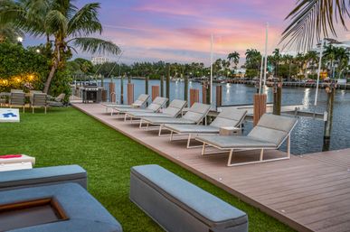 Tropical waterfront marina at sunset with modern chaise lounges on a wooden dock, palm trees, outdoor seating, grill and boats along a calm canal.