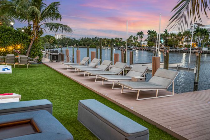 Tropical waterfront marina at sunset with modern chaise lounges on a wooden dock, palm trees, outdoor seating, grill and boats along a calm canal.