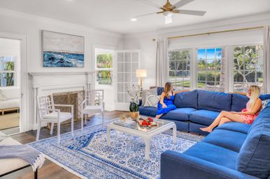 Sunlit coastal living room with a deep blue sectional, blue-and-white patterned rug, white coffee table and fireplace, large windows showing palm trees outside, and two people chatting on the sofa.