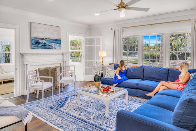 Sunlit coastal living room with a deep blue sectional, blue-and-white patterned rug, white coffee table and fireplace, large windows showing palm trees outside, and two people chatting on the sofa.