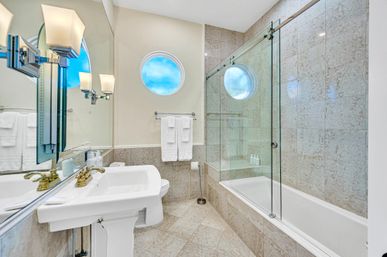 Bright modern bathroom with pedestal sink and brass faucet, glass-enclosed bathtub/shower, beige marble tile walls and floor, white towel rack and round porthole-style window.