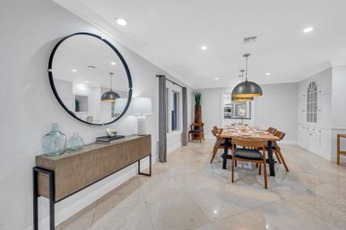 Modern open-concept dining room in a contemporary home with glossy marble floors, wooden dining table and chairs set for a meal, two large industrial pendant lights, a slim console table topped with glass vases and a round wall mirror, neutral gray-and-white palette.