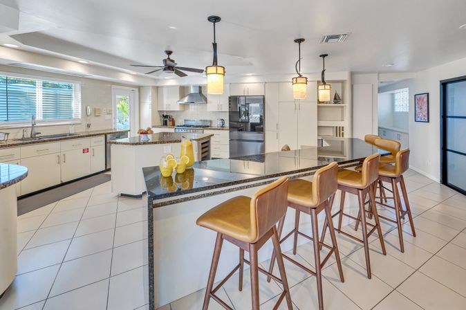 Bright modern open-plan kitchen with black granite island, four tan leather bar stools, stainless steel appliances, pendant lights, and white cabinetry.