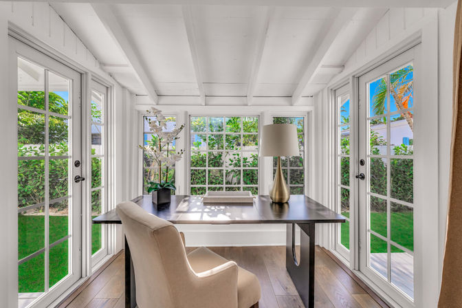 Sun-drenched sunroom home office with white-beamed ceiling, French doors and windows, dark wooden desk with brass lamp and orchid, beige upholstered chair and lush green garden view.