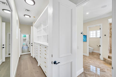 Bright walk-in closet with white built-in drawers and cubbies, hardwood floors, full-length mirror and open door leading to a marble-tiled ensuite bathroom.