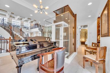 Bright luxury home foyer with a glossy black grand piano, glass elevator, sweeping curved staircase with wooden balustrades, mezzanine balcony and modern chandelier