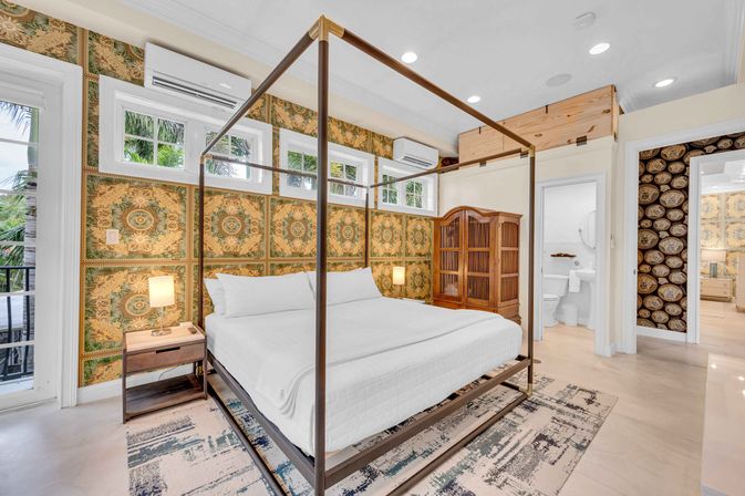Bright tropical-style bedroom with metal four-poster bed in white linens, ornate green-gold wallpaper, balcony view of palm trees, wooden armoire, nightstand lamp and en-suite bathroom