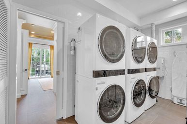 Bright laundry room with three stacked front-load washers and dryers against white subway-tiled walls, ironing board, tiled floor, and open doorway leading to a sunlit balcony with yellow curtains.