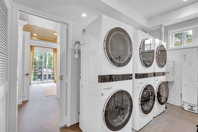 Bright laundry room with three stacked front-load washers and dryers against white subway-tiled walls, ironing board, tiled floor, and open doorway leading to a sunlit balcony with yellow curtains.