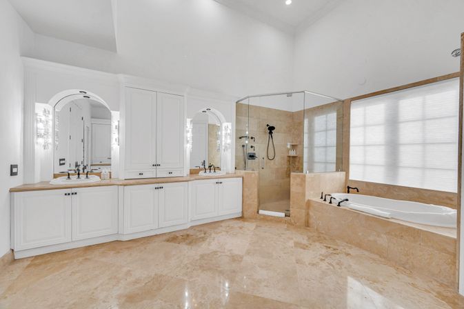 Bright spa-like luxury master bathroom with white cabinetry double vanity, marble floors, glass-enclosed shower, and built-in soaking tub beneath a large frosted window.
