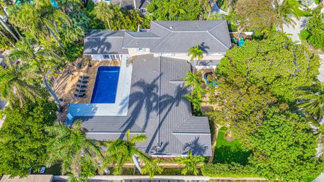 Aerial view of a South Florida tropical home: gray-roofed house, rectangular blue swimming pool with lounge chairs, and tall palm trees amid lush green landscaping.