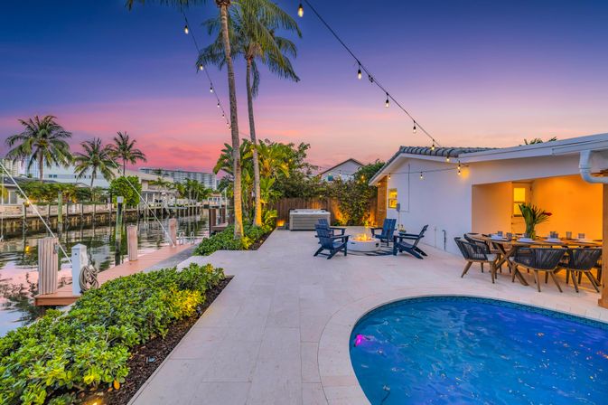 Tropical waterfront backyard at sunset with palm trees and canal-side dock, string lights, curved blue pool, outdoor dining, fire-pit seating and hot tub — coastal vacation vibe