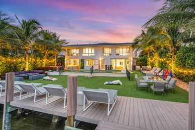 Sunset-lit waterfront modern two-story home with dock and lounge chairs, manicured lawn and pool, outdoor dining and lounge areas, palm trees, string lights and cornhole boards.