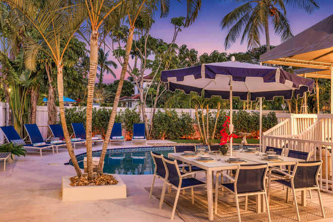 Tropical backyard poolside patio at sunset with palm trees, navy lounge chairs and an umbrella-shaded dining table set for six