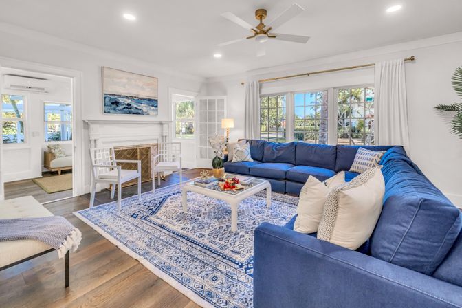 Sunny coastal-inspired living room with large windows, deep blue L-shaped sectional, white coffee table on a patterned blue area rug, white fireplace, hardwood floors and ceiling fan.