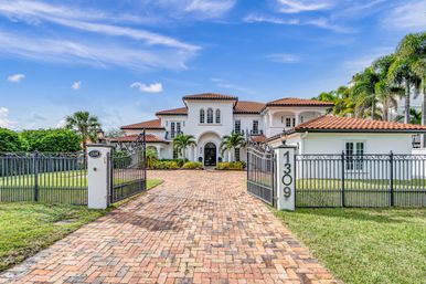 Gated Mediterranean-style luxury home with terracotta roof, arched entry, palm trees, ornamental iron gates marked 1309 and a brick paver driveway under a bright blue sky