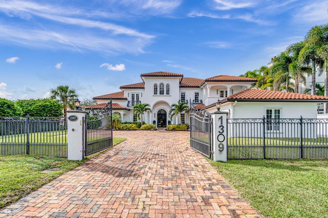 Gated Mediterranean-style luxury home with terracotta roof, arched entry, palm trees, ornamental iron gates marked 1309 and a brick paver driveway under a bright blue sky