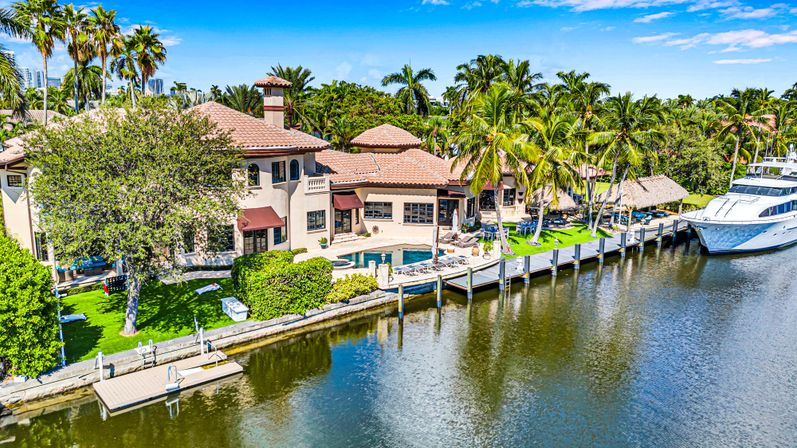 Aerial view of a Mediterranean-style waterfront villa with pool, palm trees, private dock and yacht on a sunny tropical canal.