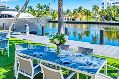 Waterfront outdoor dining at a tropical marina: a set table with blue patterned plates, glassware and white hydrangeas, palm trees, docked yacht and calm water reflections.
