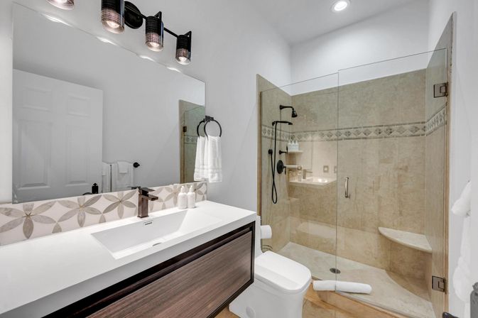 Spa-like modern bathroom with white countertop and dark wood vanity, rectangular sink and matte-black faucet, large mirror, glass-enclosed beige-tiled walk-in shower with bench and black rain/handheld shower, white toilet and folded towels.
