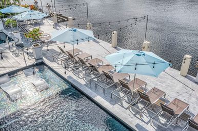 Aerial view of a chic waterfront pool deck with rows of lounge chairs, light-blue umbrellas, in-pool loungers and string lights along calm river water.