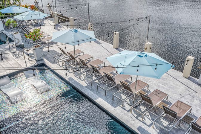 Aerial view of a chic waterfront pool deck with rows of lounge chairs, light-blue umbrellas, in-pool loungers and string lights along calm river water.