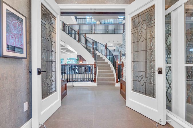 Stained-glass French doors opening to a bright multi-level grand foyer with a sweeping curved staircase, dark iron balusters, and wooden newel posts.