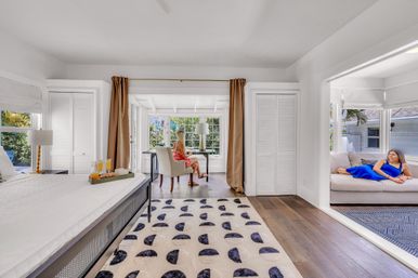 Sunlit coastal bedroom suite in a beach-house style rental: king bed with breakfast tray, patterned rug and hardwood floors, sunroom desk overlooking tropical garden with a seated person, and adjacent lounge with another person relaxing on a sofa.