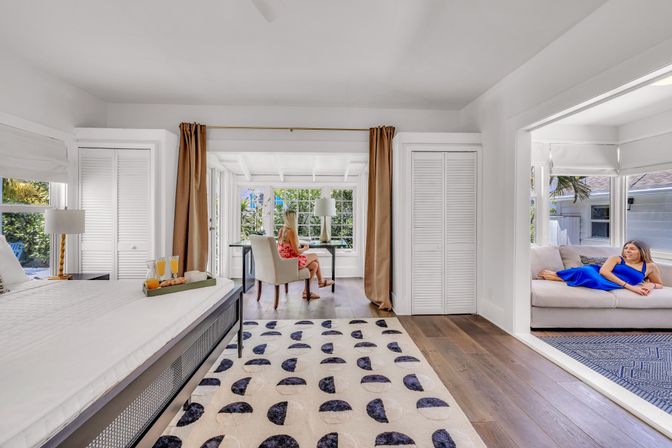 Sunlit coastal bedroom suite in a beach-house style rental: king bed with breakfast tray, patterned rug and hardwood floors, sunroom desk overlooking tropical garden with a seated person, and adjacent lounge with another person relaxing on a sofa.