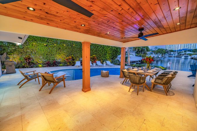 Waterfront covered patio with wooden ceiling and fan, poolside lounge chairs, outdoor dining set, and boat dock framed by tropical landscaping