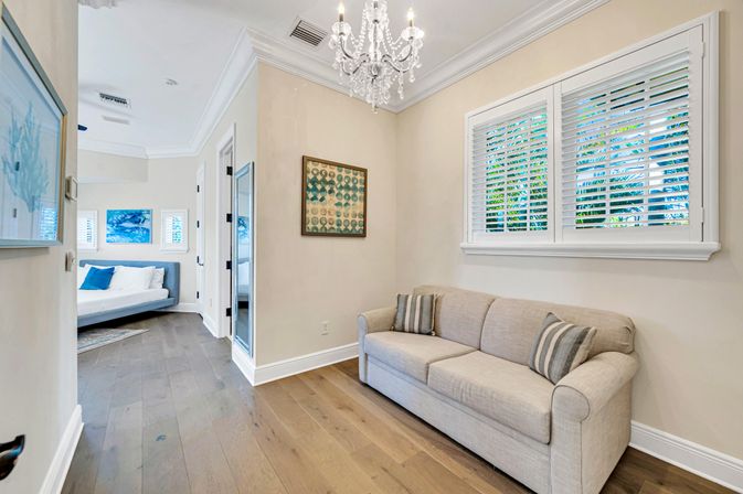 Bright coastal-style living room with beige sofa and striped pillows, crystal chandelier, white plantation shutters revealing tropical greenery, hardwood floors, and a view into a blue-accented bedroom.