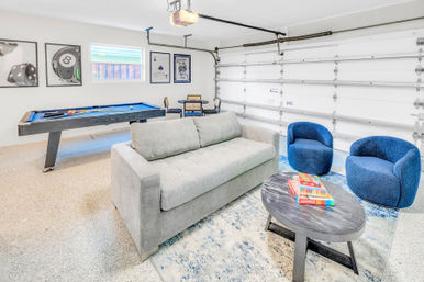 Bright, fun residential garage game room with blue-felt pool table, light gray sofa, two blue lounge chairs, round coffee table with board games, framed wall art and closed white garage door.