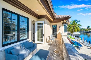 Sunny tropical waterfront balcony with blue outdoor sofa and chairs, tiled roof and French doors opening to a terrace overlooking a pool, palm trees, dock and yacht along a calm canal.