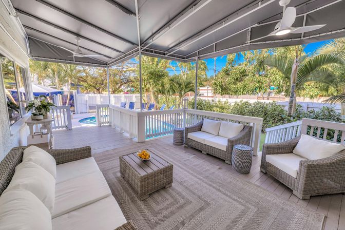 Covered tropical backyard deck with wicker sofas and cushions, rattan coffee table with fruit, ceiling fans, and railing overlooking a fenced yard and blue swimming pool surrounded by palm trees