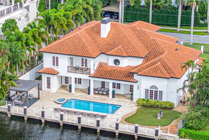 Aerial view of a Mediterranean-style waterfront villa with terracotta tile roof, private pool and sun deck, dock, and lush palm-tree landscaping.
