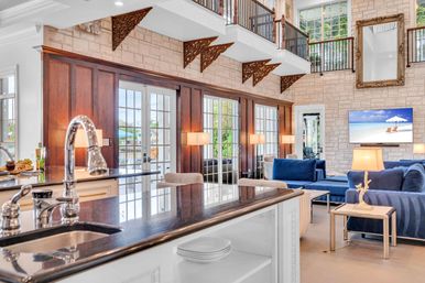 Bright open-plan coastal living room and kitchen with dark stone island and stainless sink, blue sofas, stone accent wall, tall French doors and second-story balcony