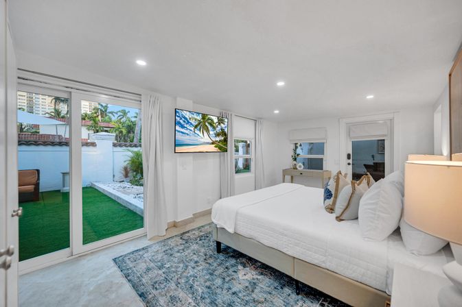 Bright coastal-style bedroom with king bed, decorative pillows and area rug, wall-mounted TV, and sliding glass doors opening to a small tropical patio with artificial turf and palm trees.