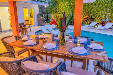 Tropical backyard patio at dusk with wooden dining table set for six with blue plates and glasses, vase of red tropical flowers centerpiece, poolside lounge chairs and grill in the background.