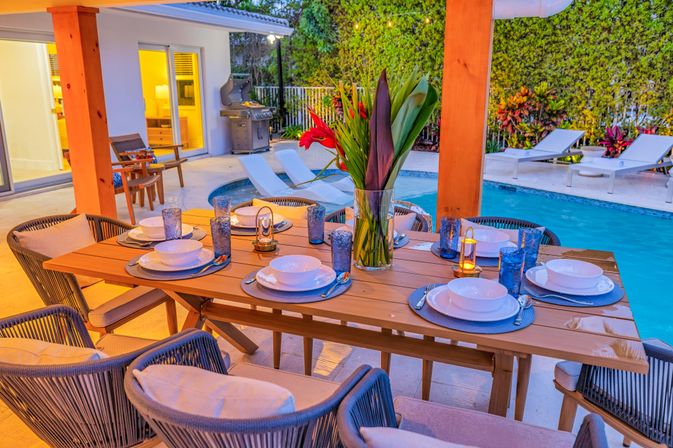 Tropical backyard patio at dusk with wooden dining table set for six with blue plates and glasses, vase of red tropical flowers centerpiece, poolside lounge chairs and grill in the background.