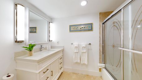 Bright modern white bathroom with single marble vanity, large mirror flanked by vertical lights, potted plant, towel rack with white towels, and frosted glass shower doors over beige tile.