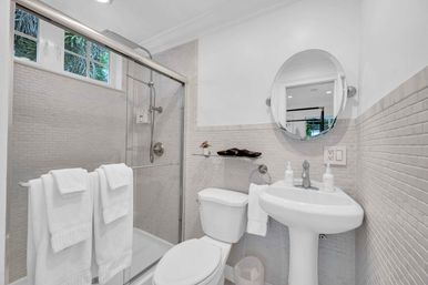 Bright, spa-like modern bathroom with glass-enclosed shower, white towels on a rail, pedestal sink, round mirror and neutral mosaic tile walls