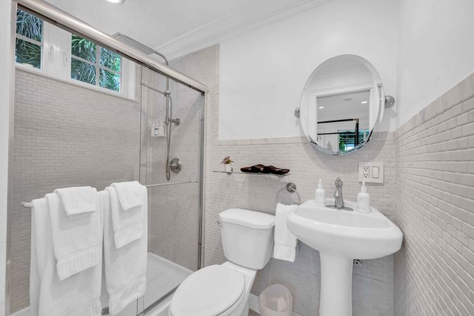 Bright, spa-like modern bathroom with glass-enclosed shower, white towels on a rail, pedestal sink, round mirror and neutral mosaic tile walls