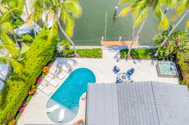 Aerial view of a tropical canal-front backyard with a kidney-shaped pool, sun loungers, palm trees, concrete patio with fire-pit seating, hot tub, and a private dock on calm water.