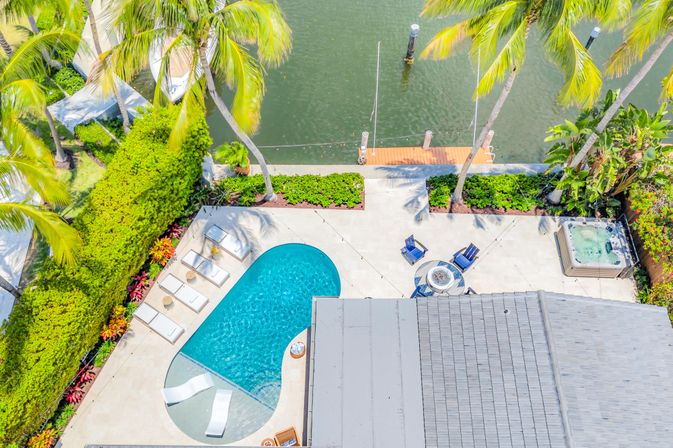 Aerial view of a tropical canal-front backyard with a kidney-shaped pool, sun loungers, palm trees, concrete patio with fire-pit seating, hot tub, and a private dock on calm water.