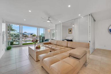 Sunlit coastal waterfront condo living room with large tan leather sectional, modern coffee table and floor-to-ceiling glass doors revealing marina, dock and palm trees.