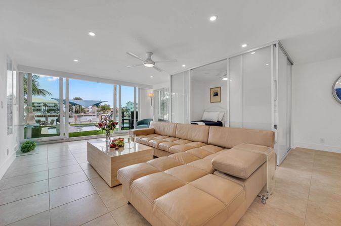 Sunlit coastal waterfront condo living room with large tan leather sectional, modern coffee table and floor-to-ceiling glass doors revealing marina, dock and palm trees.
