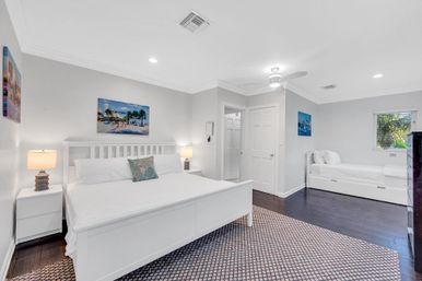 Bright coastal-style bedroom with white king bed and matching daybed, dark hardwood floors, patterned area rug, ceiling fan, bedside lamps, tropical artwork and a window view of palm trees.