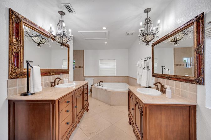 Spa-like luxury master bathroom with matching wood vanities, ornate gilt mirrors, crystal chandeliers, dual sinks, and a corner soaking tub with beige marble tile.